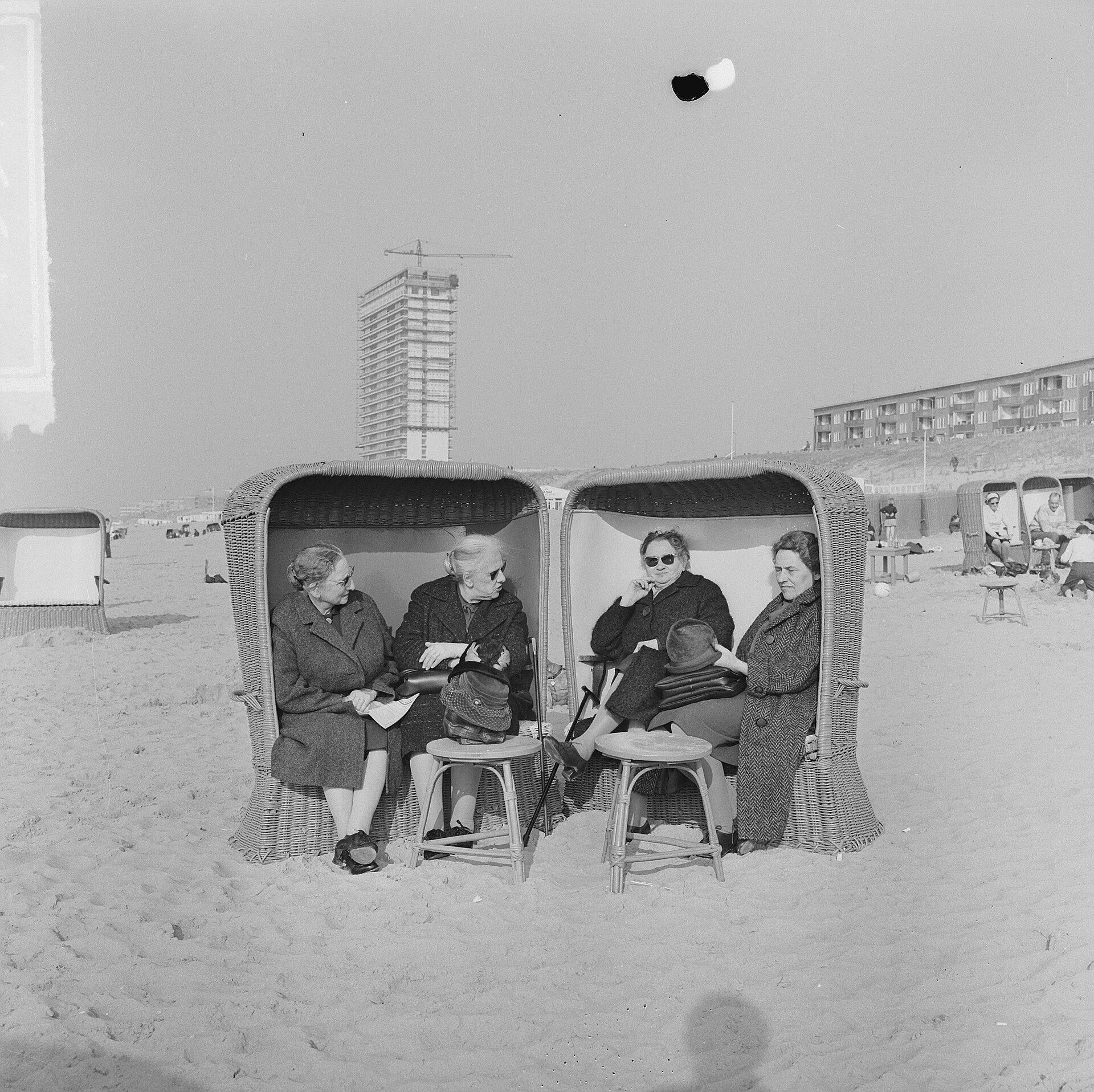 Zwart-witfoto uit 1965: vier dames in winterjas zitten in twee rieten strandstoelen op het strand van Zandvoort
