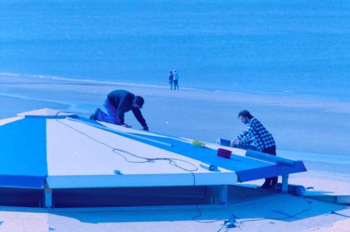 Kleurenfoto uit 2001: twee bouwvakkers leggen het dak op het paviljoen, op het strand loopt een stel langs de zee