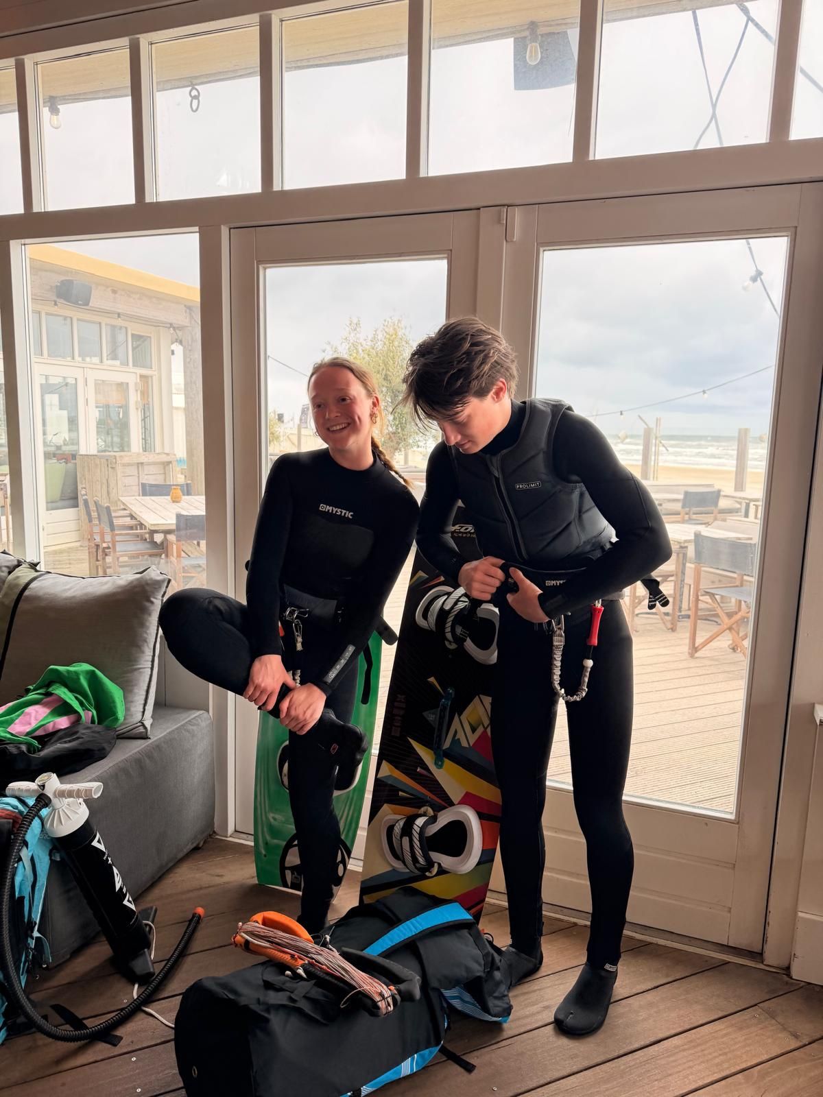 Twee kiters in wetsuit maken hun spullen klaar bij de ramen van NIUS, met de zee op de achtergrond