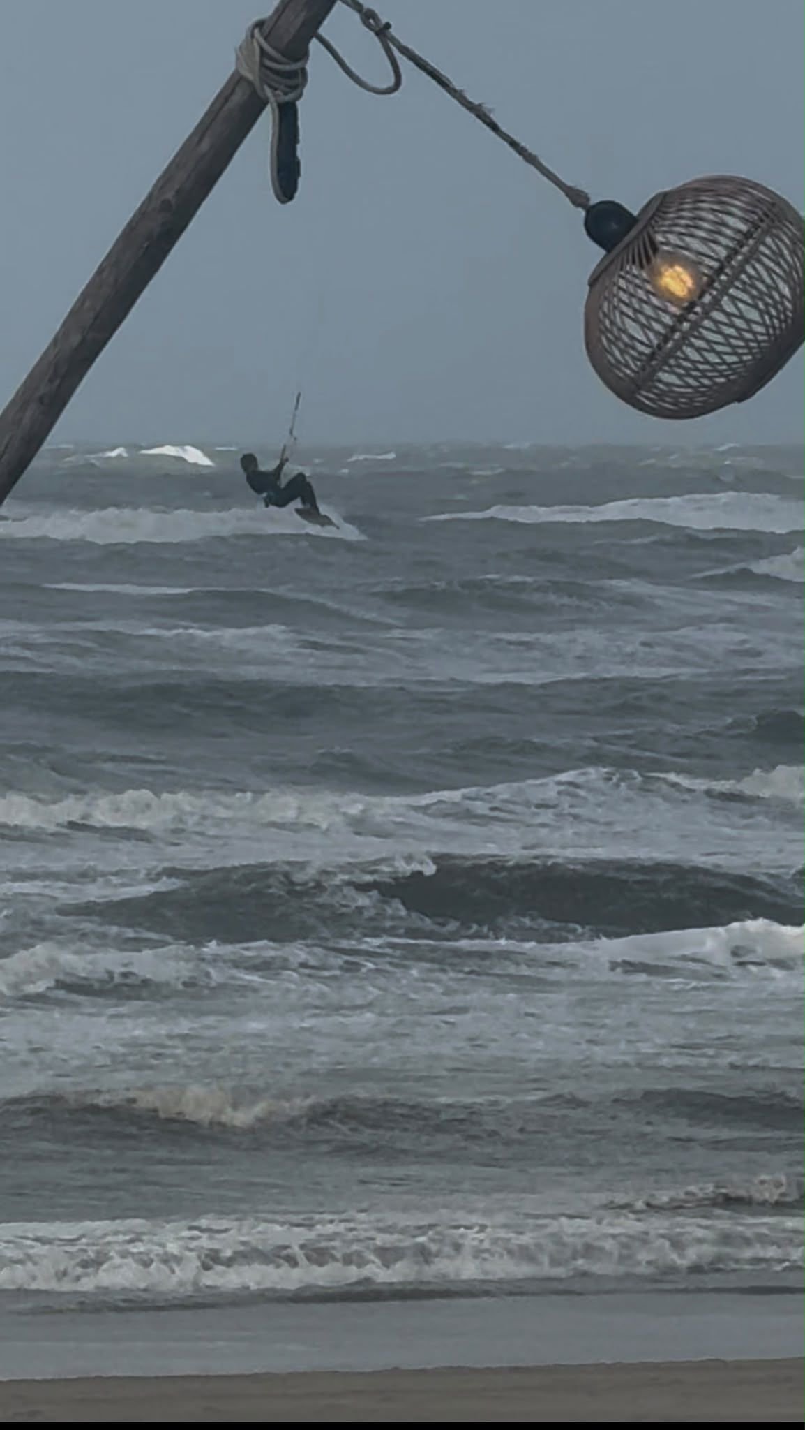 Een kiter maakt een sprong in de golven, gezien vanaf het terras van NIUS onder een brandende rieten lamp