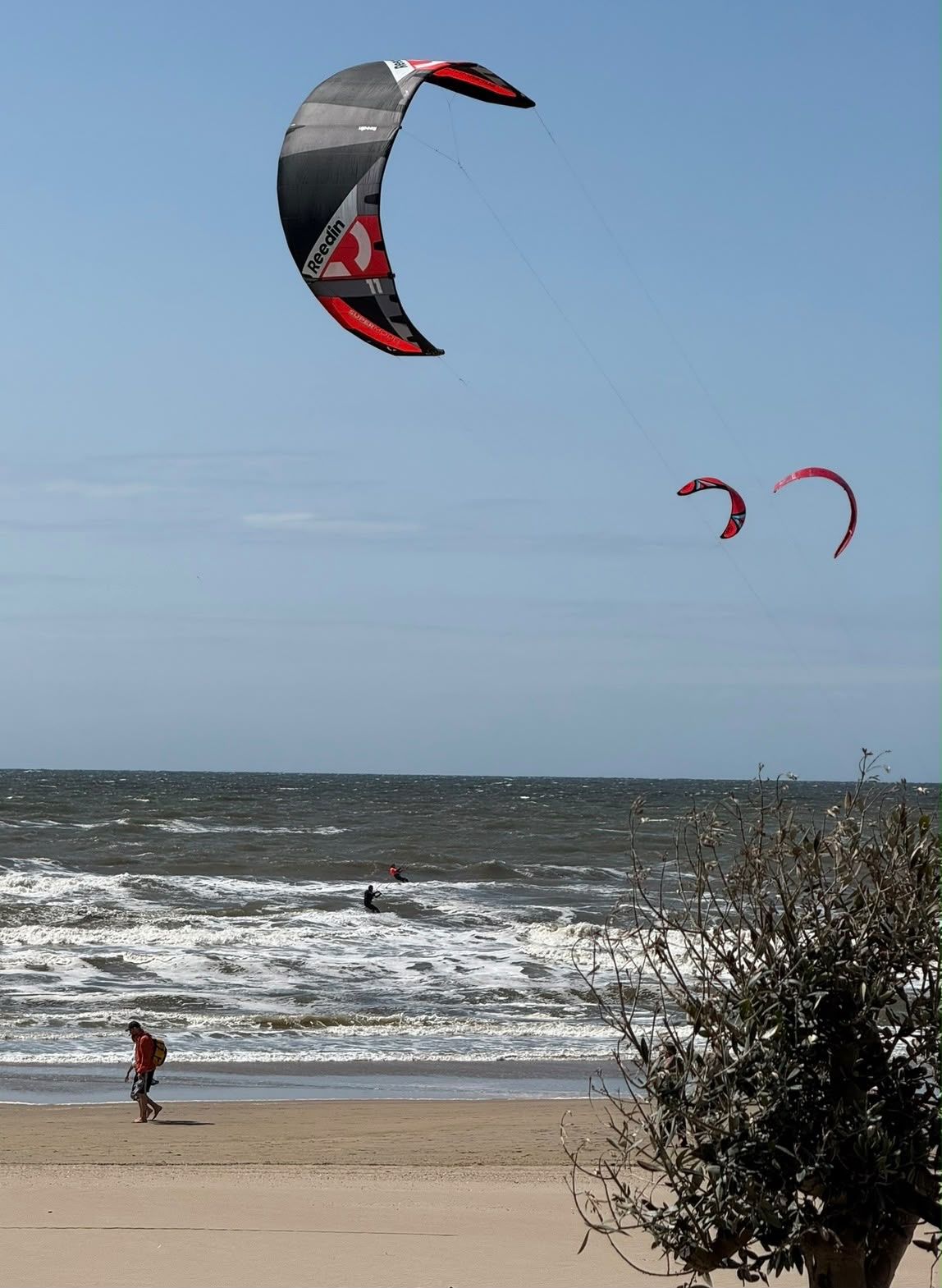 Drie kites in de lucht boven de zee bij Zandvoort, met twee kiters in de branding en een wandelaar op het strand