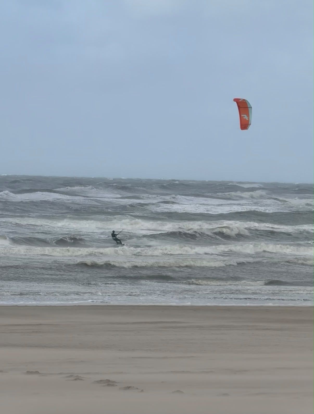Een kiter met een oranje kite in de woeste branding bij Zandvoort, met stuivend zand op het strand