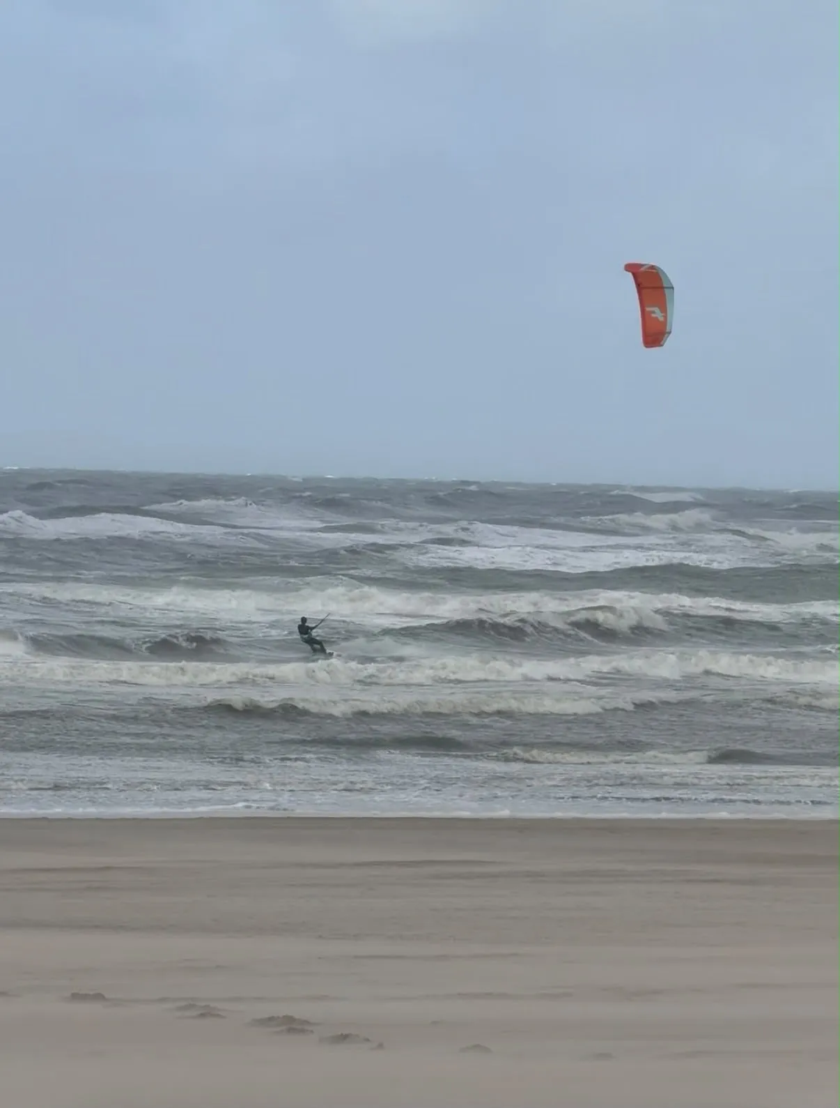 Een kiter met een oranje kite in de woeste branding bij Zandvoort