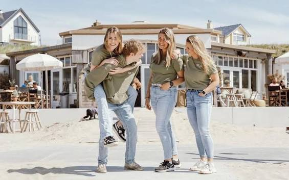 Vier collega’s in olijfgroene NIUS-shirts lachen op het strand voor het paviljoen